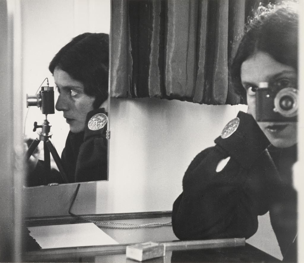 A black-and-white self-portrait by Ilse Bing showing her twice: in profile on the left behind her Leica camera on a tripod, and facing forward on the right, her eye visible above the camera lens, the composition split by mirror frames and a dark curtain.