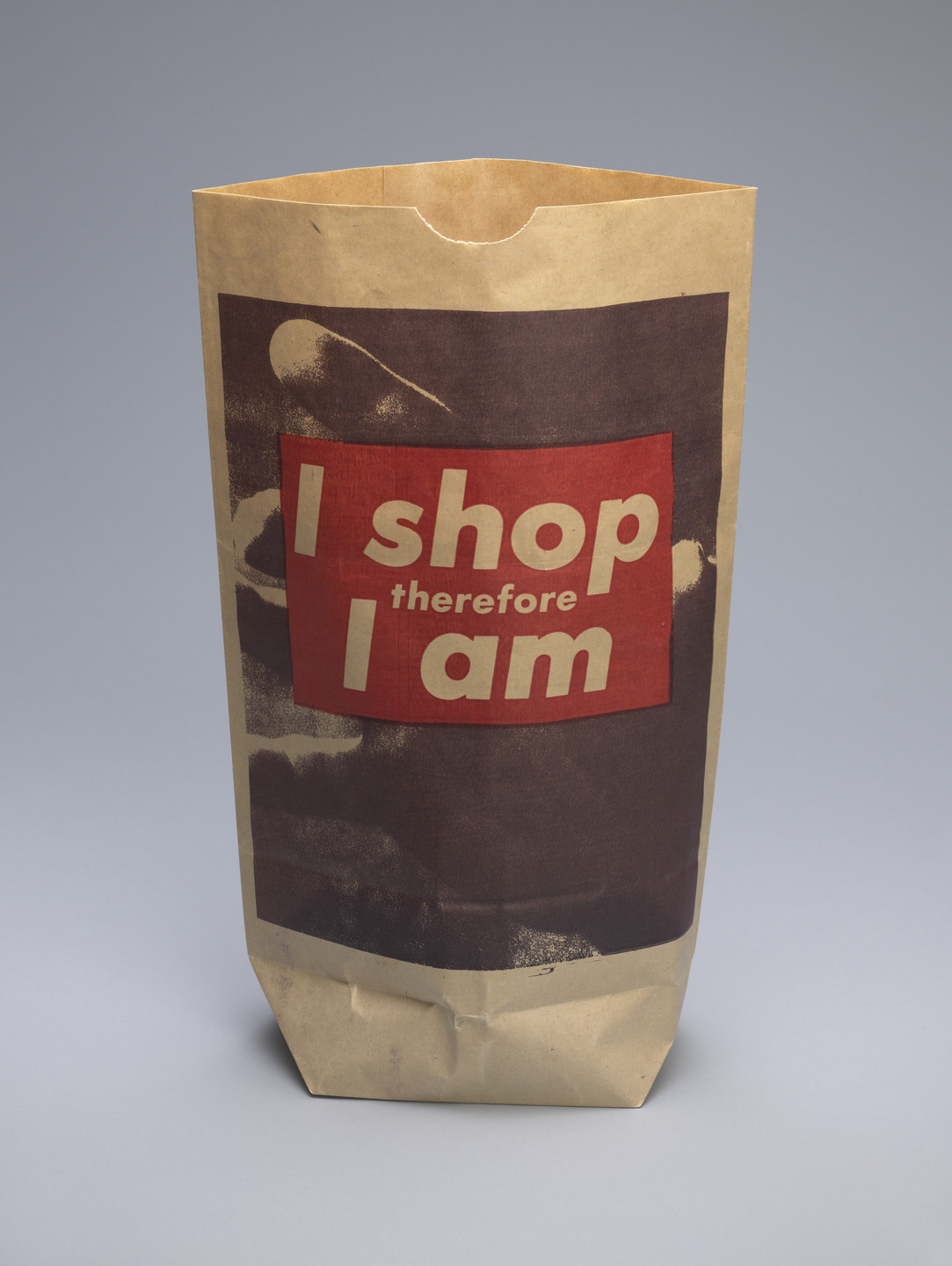 A brown paper bag with red and white text that reads, &#34;I shop therefore I am.&#34; The bag has a dark brown image behind the text. This is an AI generated description. To give feedback, email ai@moma.org.