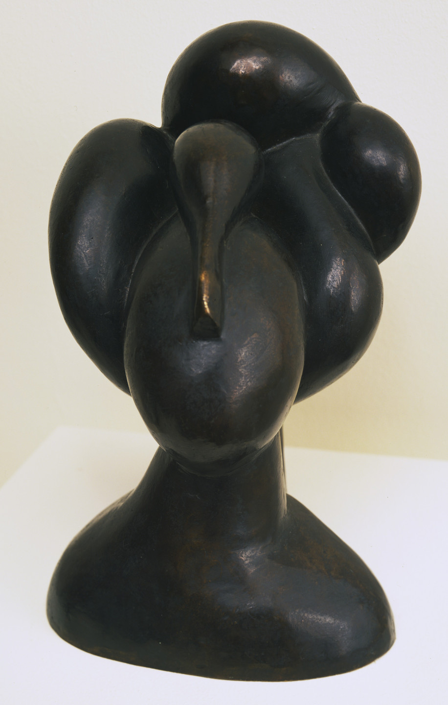 A dark bronze sculpted head with large, smooth, rounded forms suggesting hair or headgear, and an elongated central protrusion. The sculpture sits against a light background on a white surface. This is an AI generated description. To give feedback, email ai@moma.org.