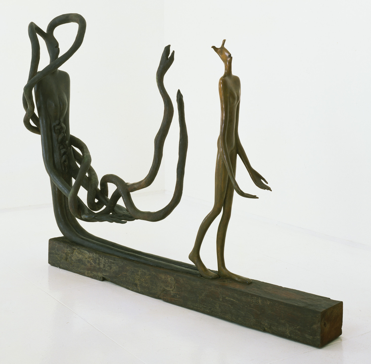An image of a bronze sculpture with two elongated humanoid figures, one with tentacle-like extensions and the other walking. The figures stand on a rectangular wooden base against a plain white background. This is an AI generated description. To give feedback, email ai@moma.org.