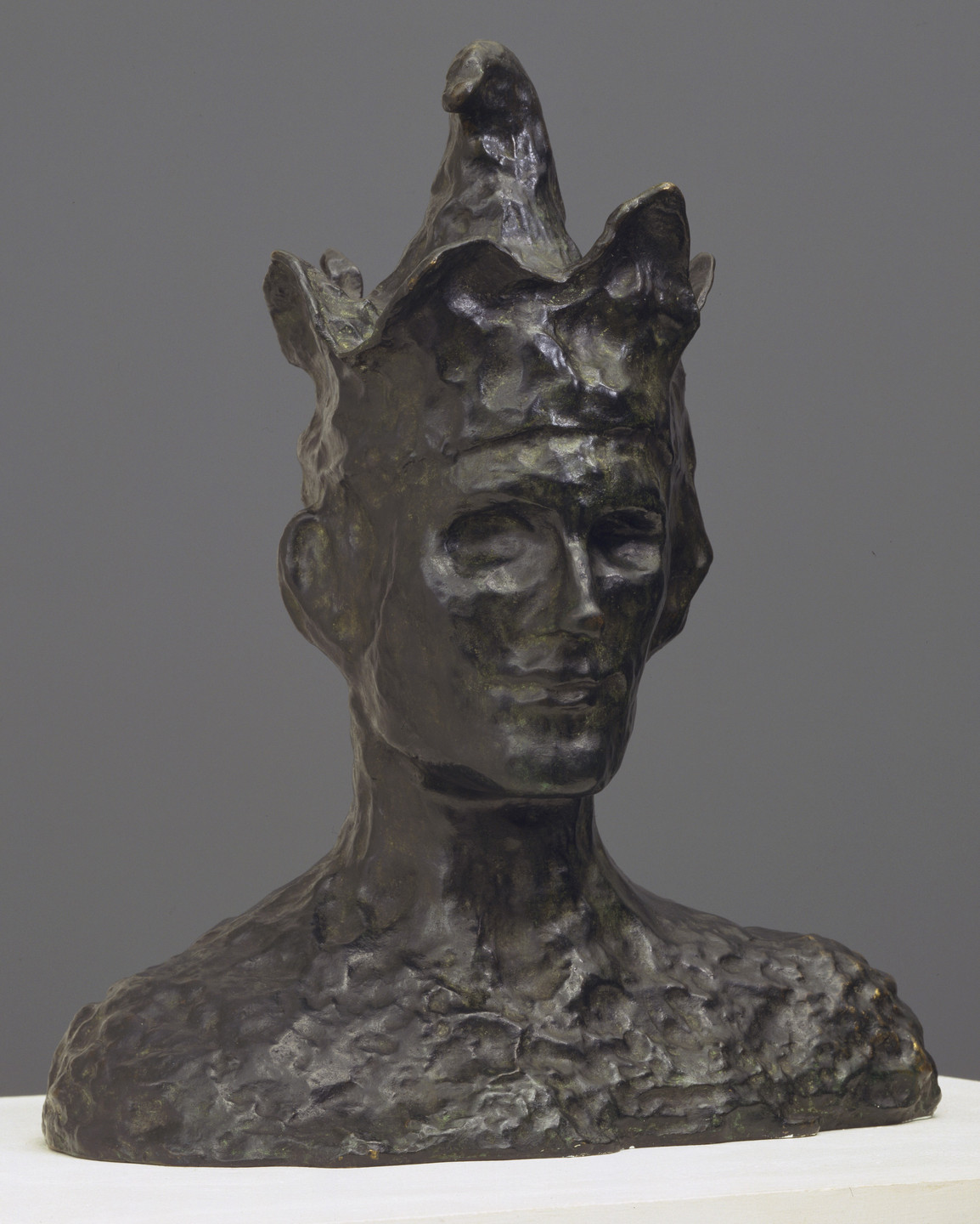 A dark bronze bust portrays a figure with closed eyes and a textured surface, wearing a crown-like headpiece. The background is plain gray, and the bust sits on a white base. This is an AI generated description. To give feedback, email ai@moma.org.