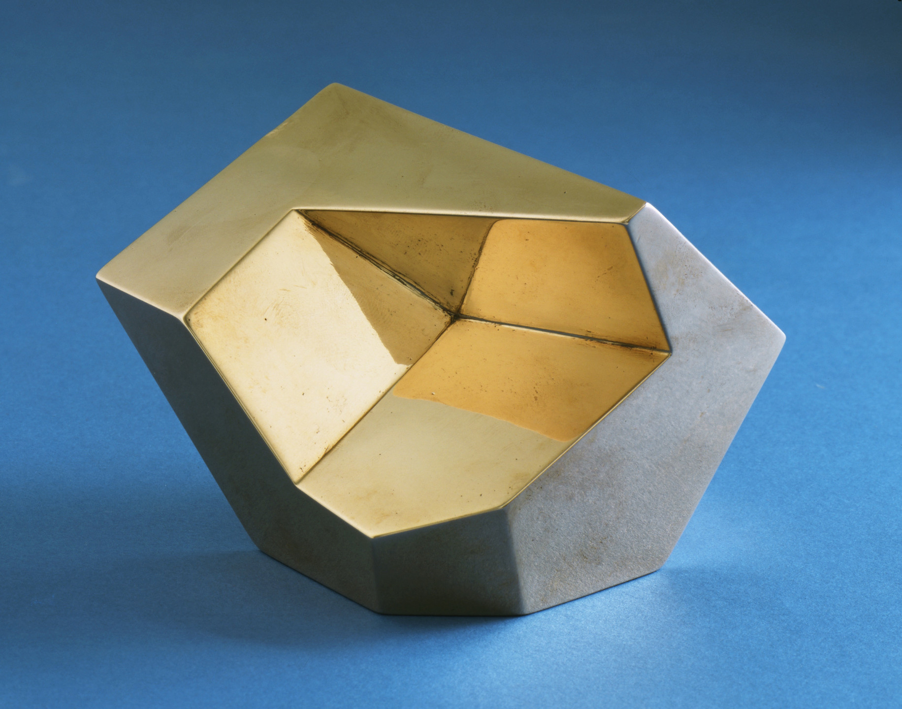 A geometric object with a mix of gold and gray surfaces is shown against a blue background. It has an angular, faceted shape with parts that reflect light. This is an AI generated description. To give feedback, email ai@moma.org.