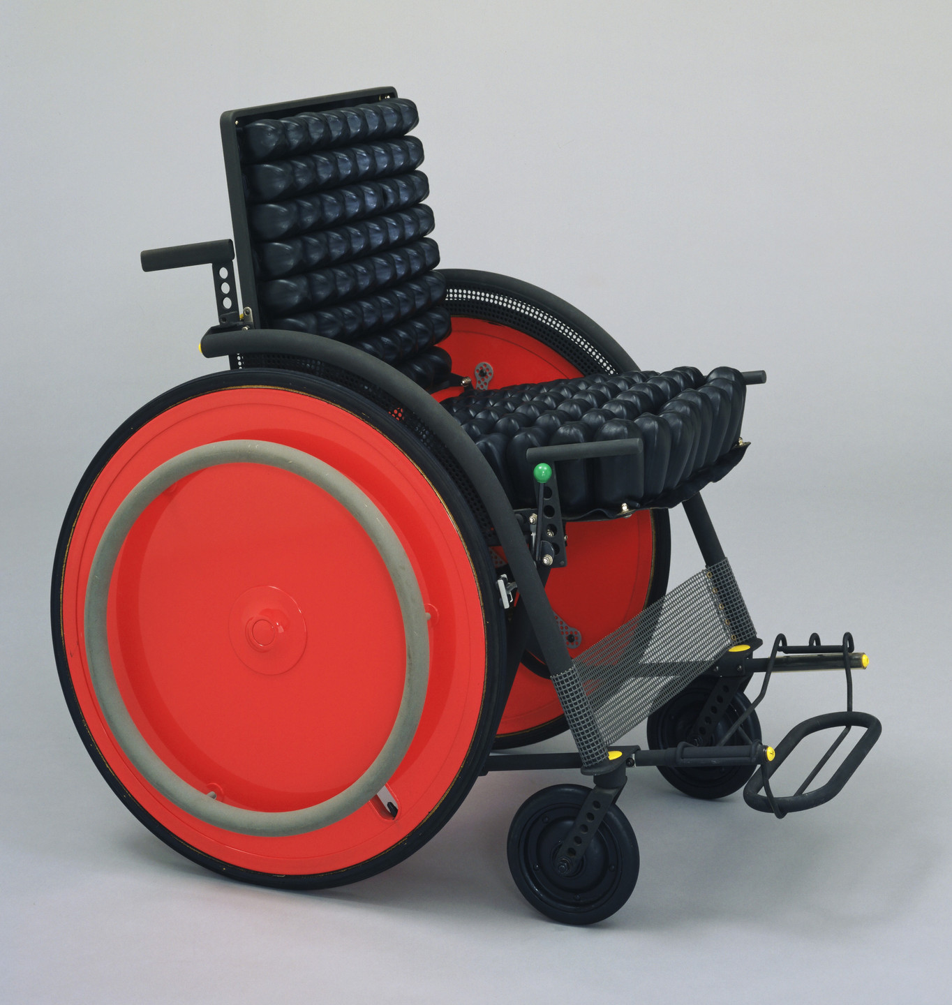 A wheelchair with a black frame, black padded seat and backrest, large bright red back wheels, and small black front wheels. The footrests are black with yellow accents. This is an AI generated description. To give feedback, email ai@moma.org.