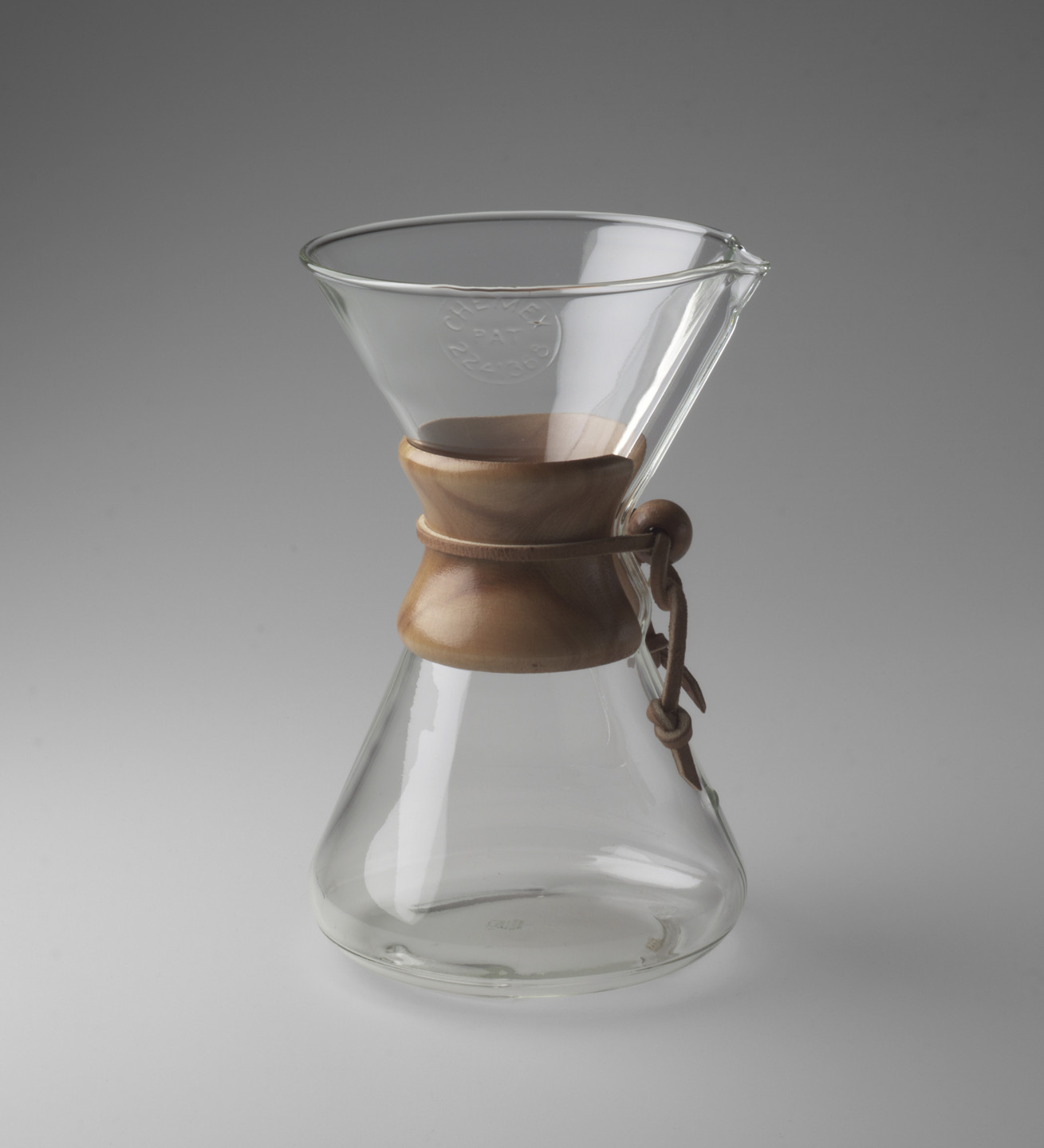 A clear, hourglass-shaped glass vessel with a wide top and narrow bottom sits on a gray background. It has a wooden collar around the middle, tied with a leather strap. This is an AI generated description. To give feedback, email ai@moma.org.