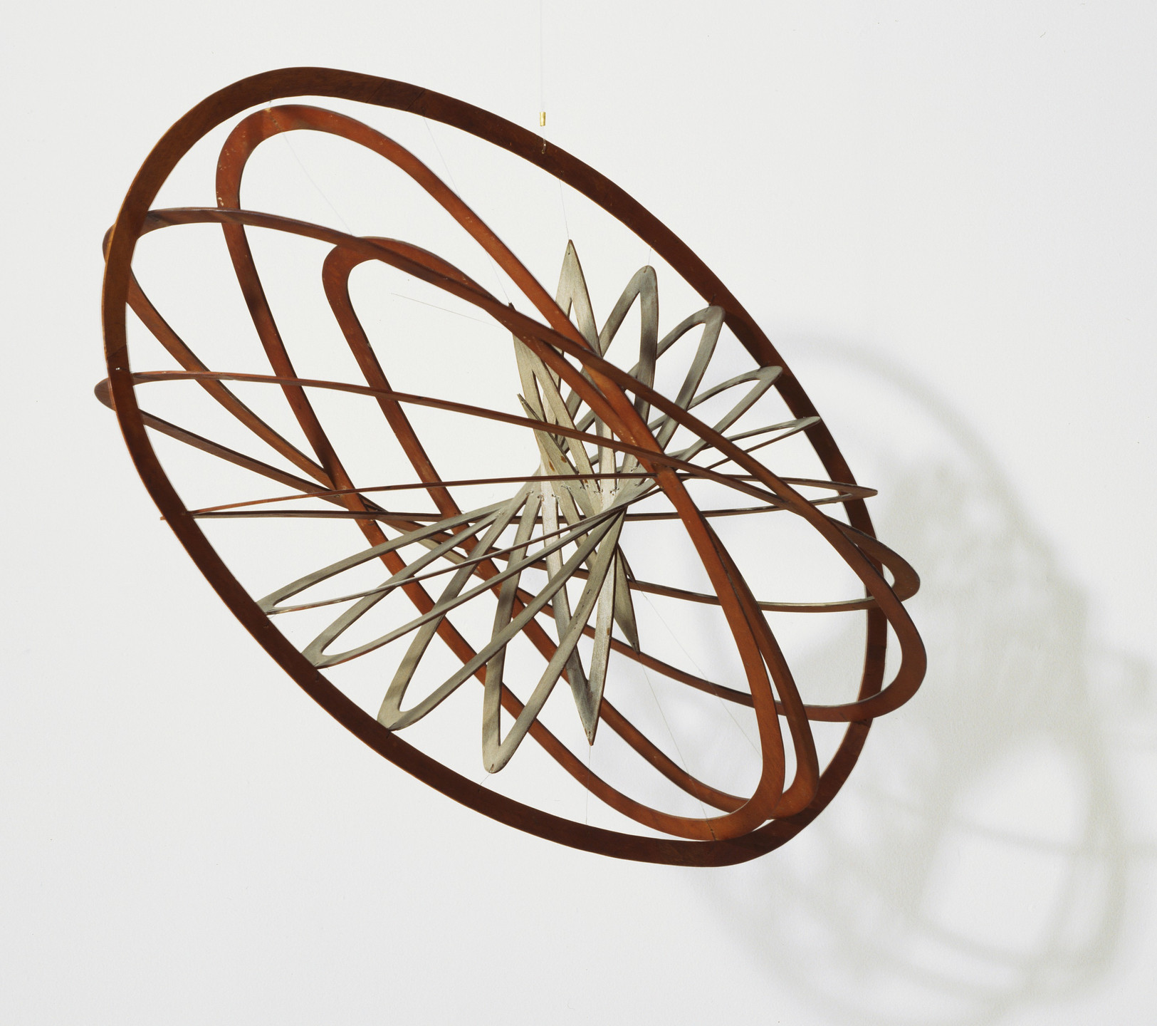 A hanging sculpture is composed of interlocking brown and silver curved strips forming an intricate, spherical design. The background is a plain white wall. This is an AI generated description. To give feedback, email ai@moma.org.