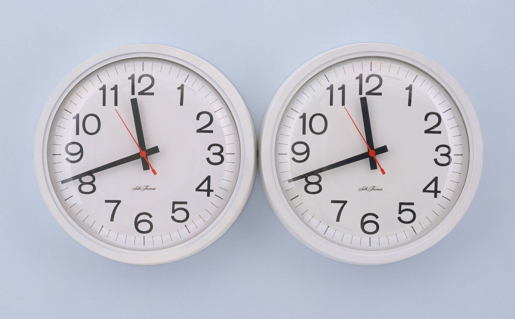 Two identical wall clocks with white faces and black numbers are set at different times against a light blue wall. The clock on the left shows 10:10, and the one on the right shows 10:11. This is an AI generated description. To give feedback, email ai@moma.org.