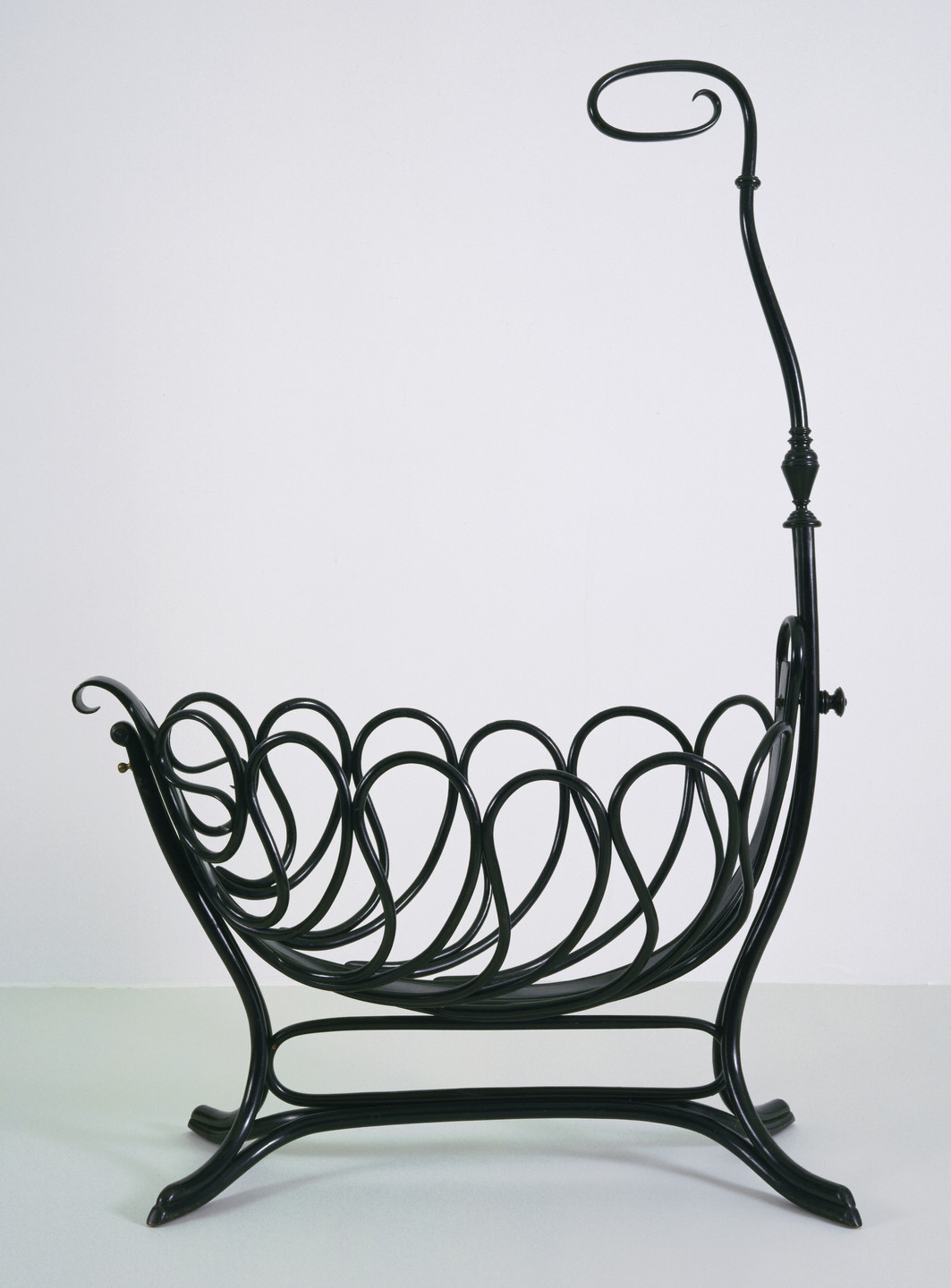A black, ornate metal magazine rack with swirling loops forms a bowl-like structure, standing on four curved legs. An additional high stand curls into a spiral at the top. This is an AI generated description. To give feedback, email ai@moma.org.