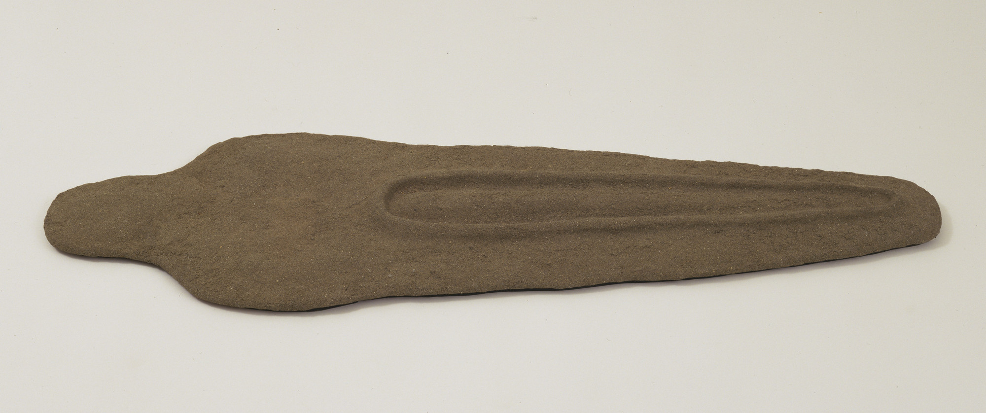 A flat, brown stone object with a roughly sword-like shape lies on a plain white background. The stone has a groove running lengthwise down its center. This is an AI generated description. To give feedback, email ai@moma.org.
