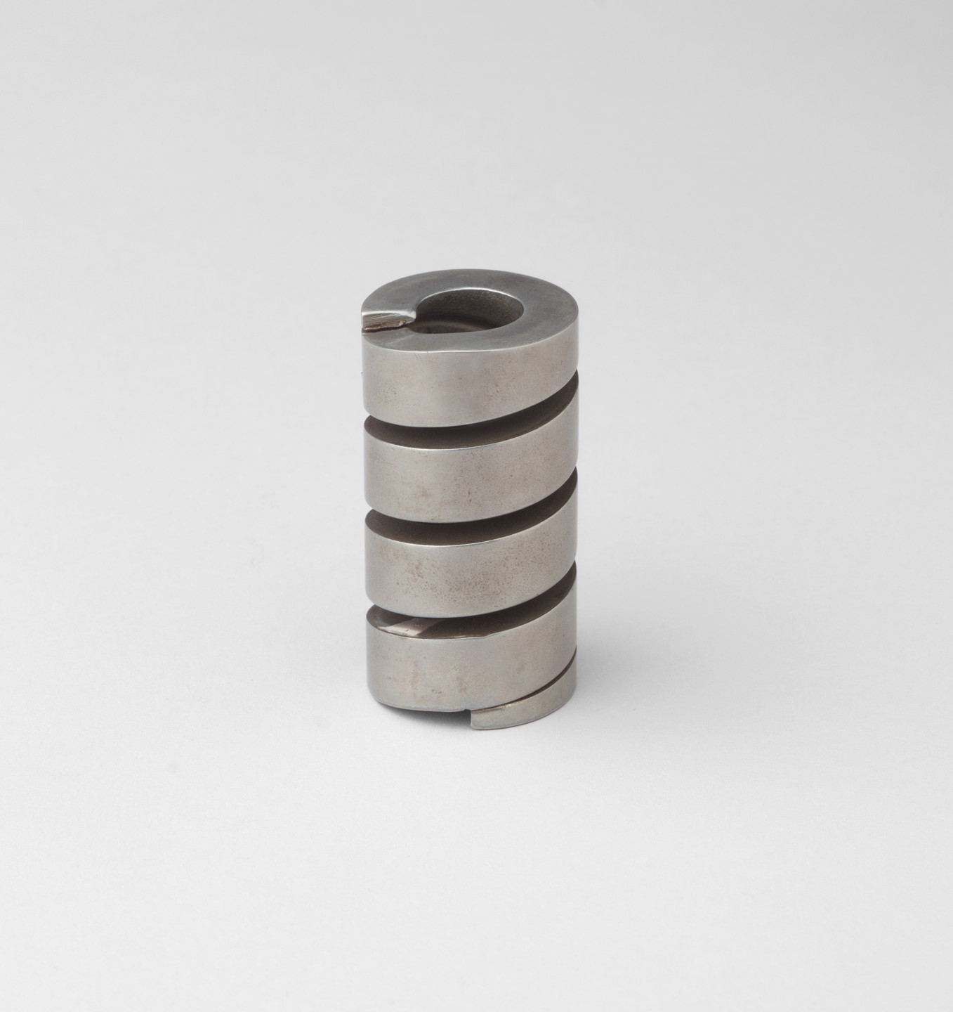 A cylindrical metallic sculpture with a grey surface is segmented into four horizontal sections with a continuous, spiraling groove. It stands upright against a plain, light grey background. This is an AI generated description. To give feedback, email ai@moma.org.