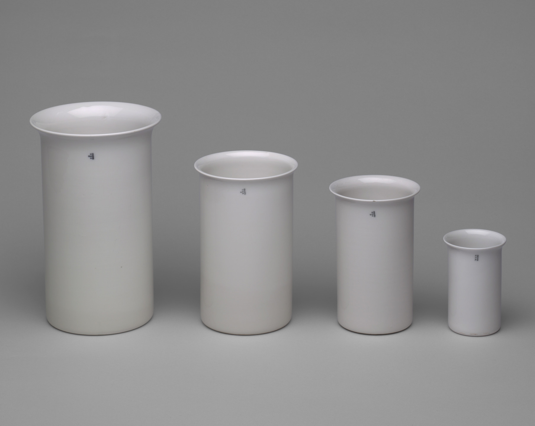 The image shows four white cylindrical containers in a row from left to right, decreasing in size. Each container has a small, dark number "4" near the top. This is an AI generated description. To give feedback, email ai@moma.org.
