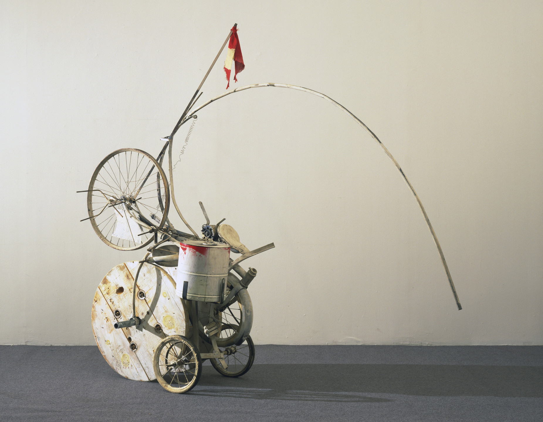 A sculpture composed of various objects, including a bicycle wheel, metal barrel, curved rods, and a red and yellow flag. The background is beige, and the floor is dark gray. This is an AI generated description. To give feedback, email ai@moma.org.