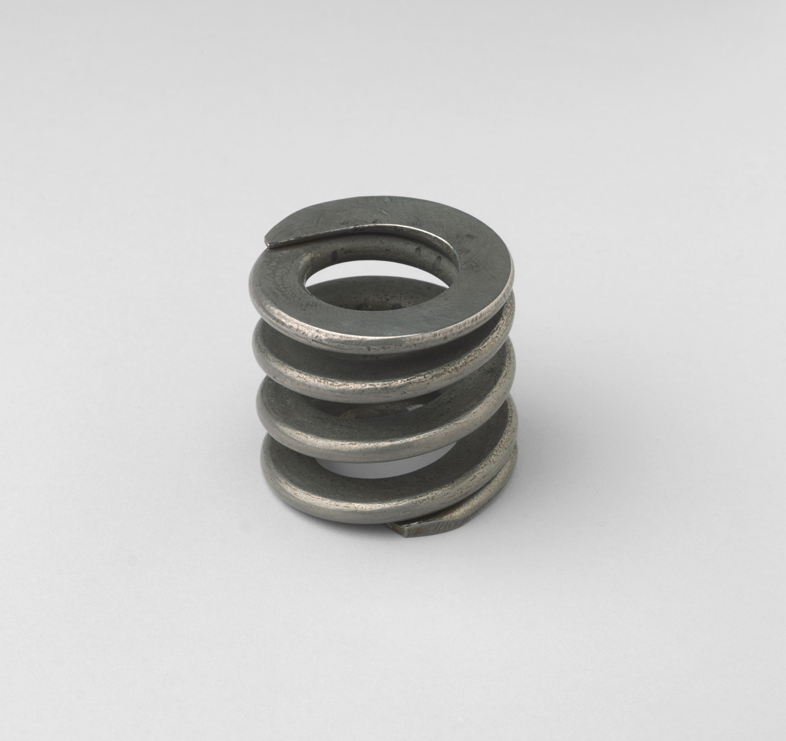 A metallic helical spring with four coils is standing upright on a plain, light-gray background. The spring has a dark silver hue and appears to be made of metal. This is an AI generated description. To give feedback, email ai@moma.org.