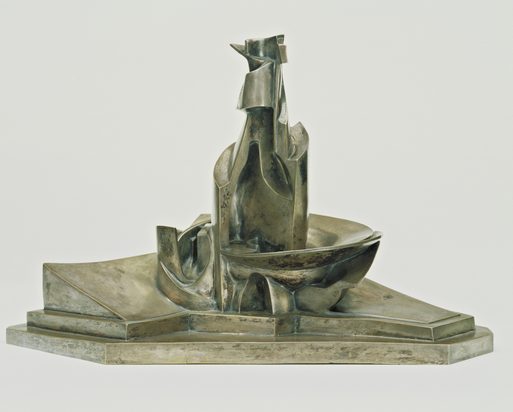 A metallic sculpture with interlocking and overlapping geometric shapes stands on a tiered, multi-sided base. The predominant color is a weathered bronze. This is an AI generated description. To give feedback, email ai@moma.org.