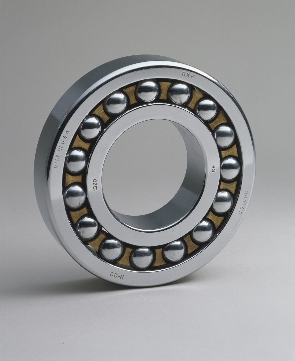 A metallic circular ball bearing with visible steel balls is shown against a plain gray background. The bearing has a silver outer ring with text, and a gold inner frame holding the balls. This is an AI generated description. To give feedback, email ai@moma.org.