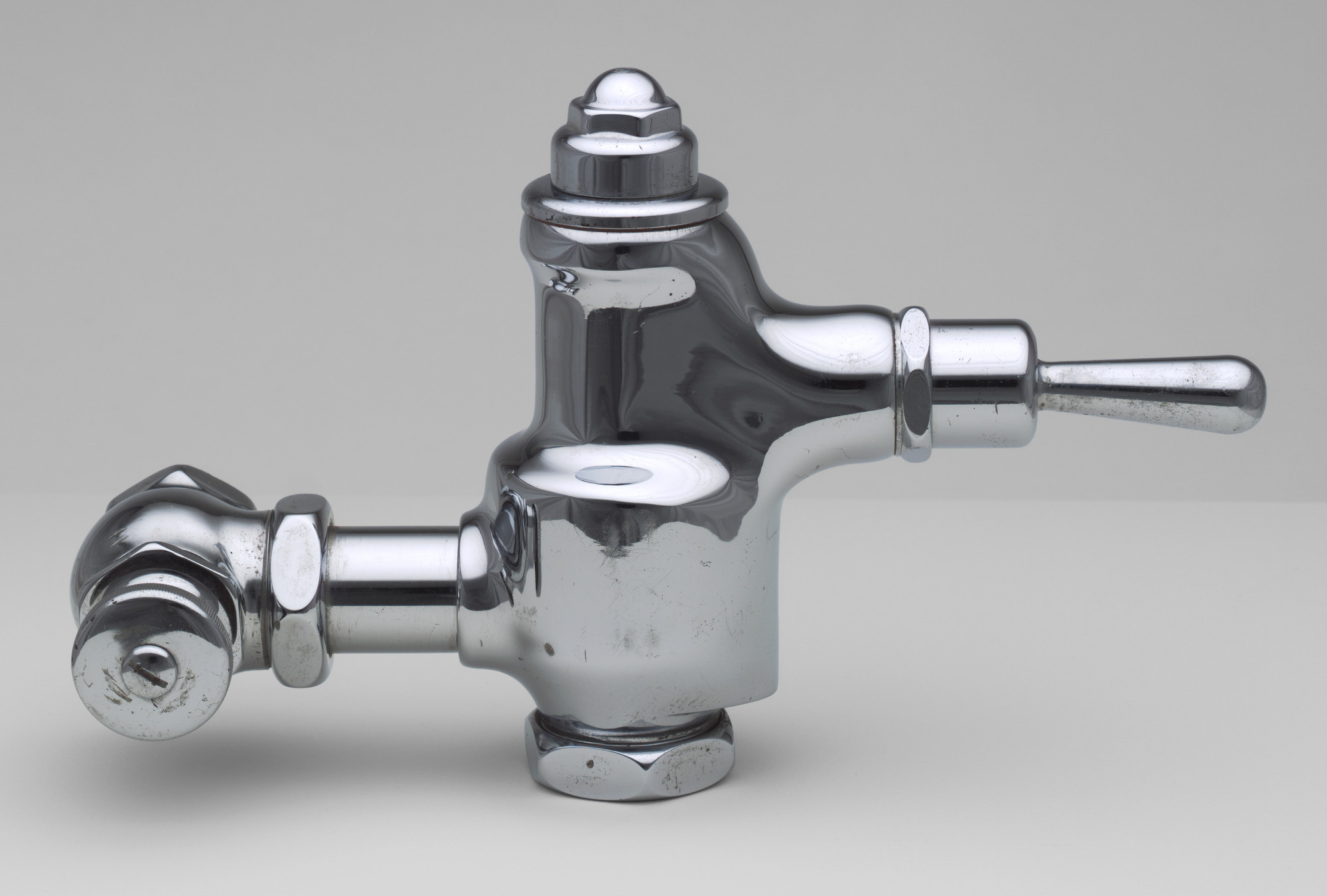 A metallic silver faucet with a cylindrical handle and a valve mechanism is set against a plain gray background. The faucet has a shiny, reflective surface with some visible smudges and scratches. This is an AI generated description. To give feedback, email ai@moma.org.