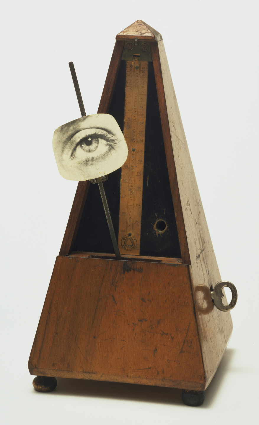 A wooden metronome with a triangular shape sits on a flat surface, featuring a mechanical arm with a black-and-white image of an eye attached to it. The metronome is predominantly brown with some metallic parts. This is an AI generated description. To give feedback, email ai@moma.org.