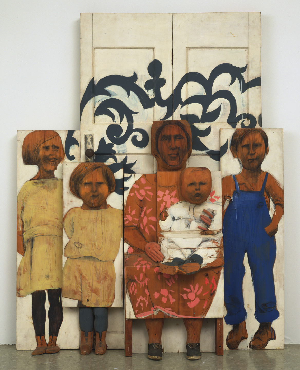 The image shows five wooden cutouts of people with painted figures. Behind the cutouts is a white, two-panel door with dark blue decorative patterns; the predominant colors are brown and blue. This is an AI generated description. To give feedback, email ai@moma.org.