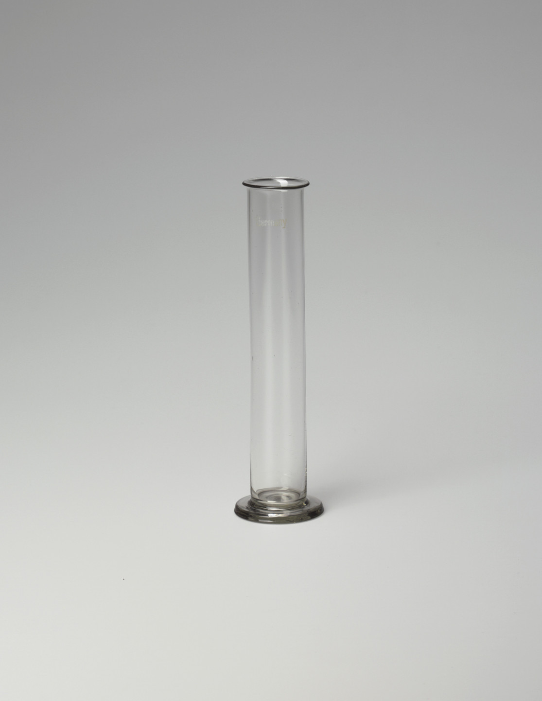A clear cylindrical glass vessel with a narrow mouth and a flat, circular base is centered against a light grey background. The vessel is empty and stands upright. This is an AI generated description. To give feedback, email ai@moma.org.