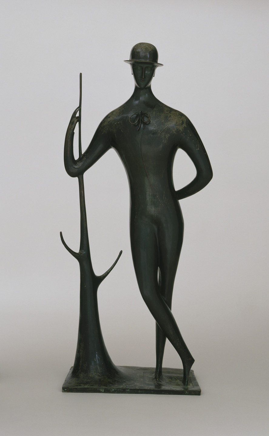 A dark green sculpture of a standing figure with elongated limbs, wearing a bowler hat, leans on a tree-like form. The figure stands on a rectangular base and has one hand on its hip. This is an AI generated description. To give feedback, email ai@moma.org.