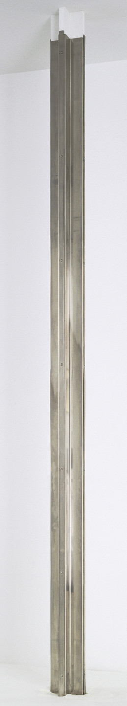 A tall, vertical metallic sculpture with symmetrical, linear ridges stands against a white background. The metallic surface has a silver-gray color and reflects light. This is an AI generated description. To give feedback, email ai@moma.org.