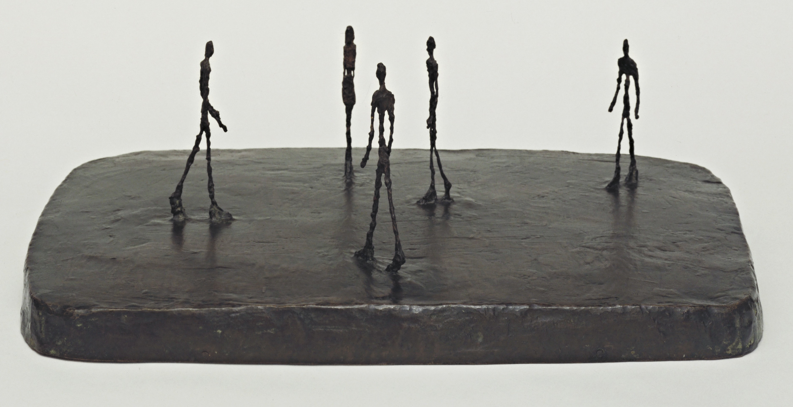 A bronze sculpture by Alberto Giacometti featuring 5 attenuated figures in walking postures on a rectangular slab.