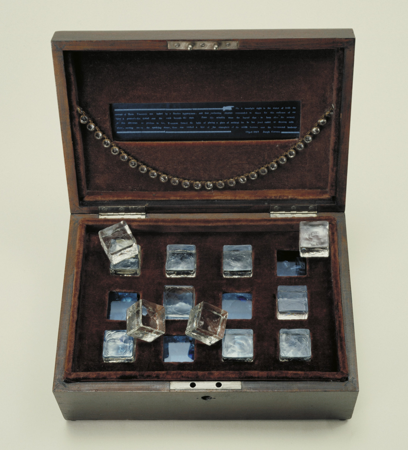 A wooden box with a dark brown velvet interior contains 15 clear glass cubes arranged in a grid pattern. The lid has a strand of metal beads and a rectangular blue panel with text on it. This is an AI generated description. To give feedback, email ai@moma.org.