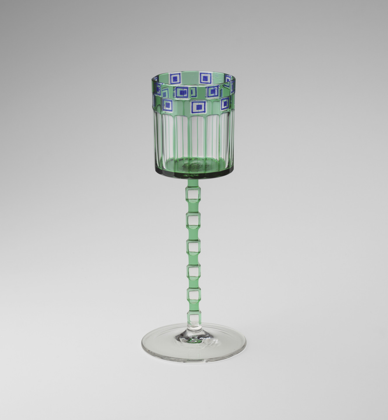 A green wine glass with a cylindrical bowl featuring blue and white square patterns near the rim. The stem consists of a series of square links and ends in a clear, flat base. This is an AI generated description. To give feedback, email ai@moma.org.