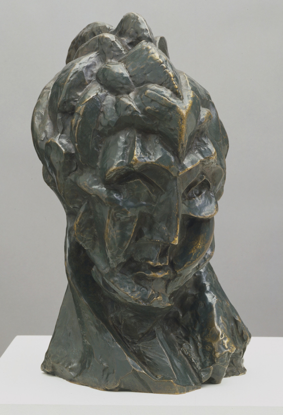 A dark green, bronze-like sculpture of a distorted human face features sharp, angular shapes and uneven surfaces. The piece is textured with gold accents and displayed on a white platform. This is an AI generated description. To give feedback, email ai@moma.org.