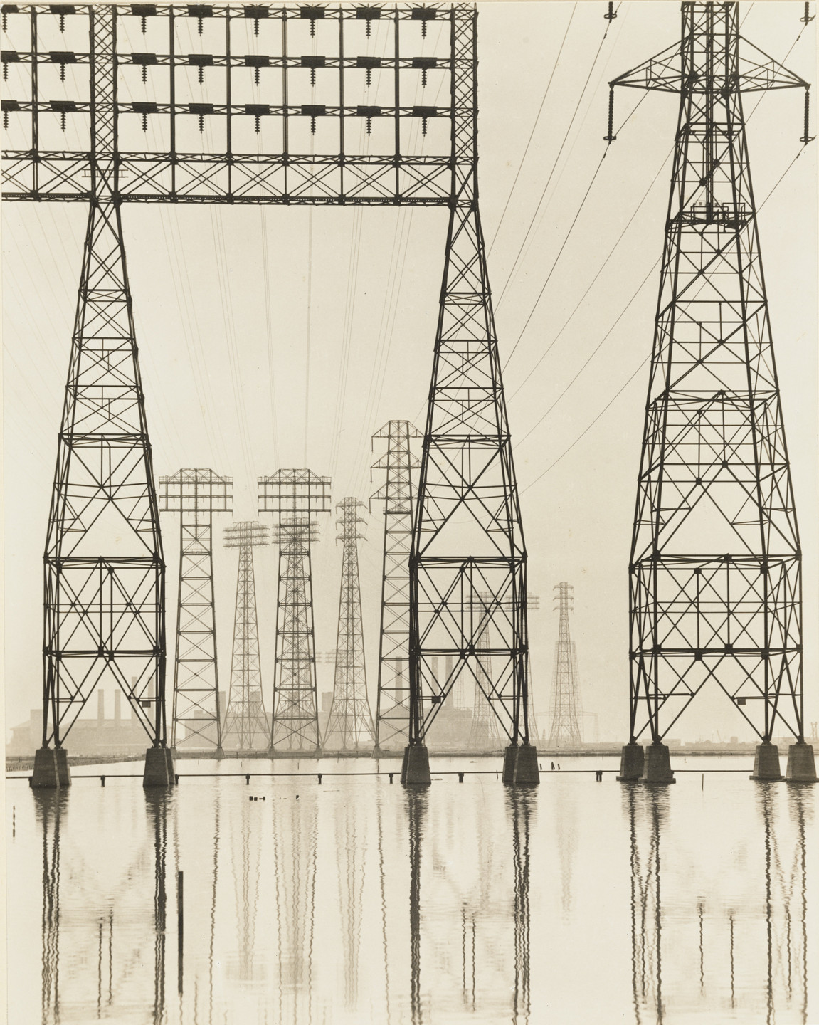 The image shows several tall, lattice steel transmission towers, reflected in a body of water, against a light gray sky. The towers are aligned in rows, receding into the background. This is an AI generated description. To give feedback, email ai@moma.org.