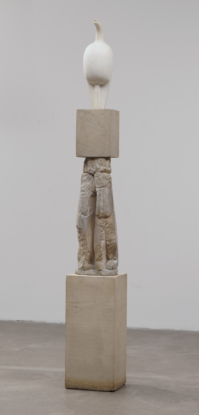 A white bird-like figure stands atop a beige stone block, which is placed on a light brown column with engraved human-like legs. The sculpture is displayed against a plain, light gray background. This is an AI generated description. To give feedback, email ai@moma.org.
