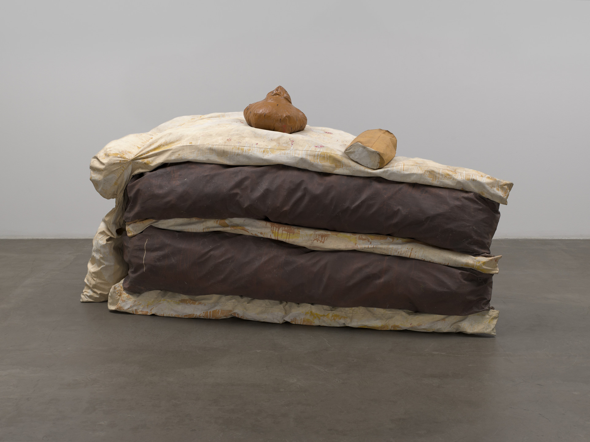 The image shows a stack of two large, unevenly folded brown cushions sandwiched between beige sheets. On top of the stack, there are two brown, organic-shaped objects resting on the beige sheet. This is an AI generated description. To give feedback, email ai@moma.org.