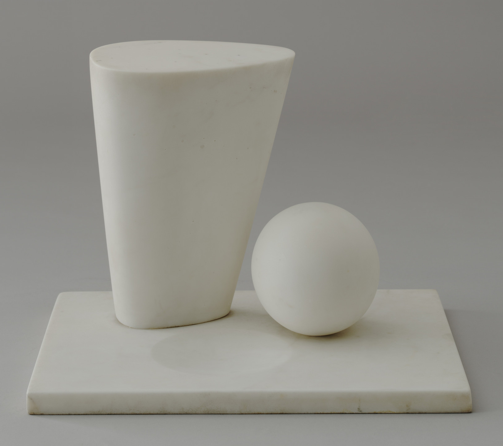 A white, uneven cylindrical shape stands upright next to a white sphere on a flat, white rectangular base. The background is solid gray. This is an AI generated description. To give feedback, email ai@moma.org.