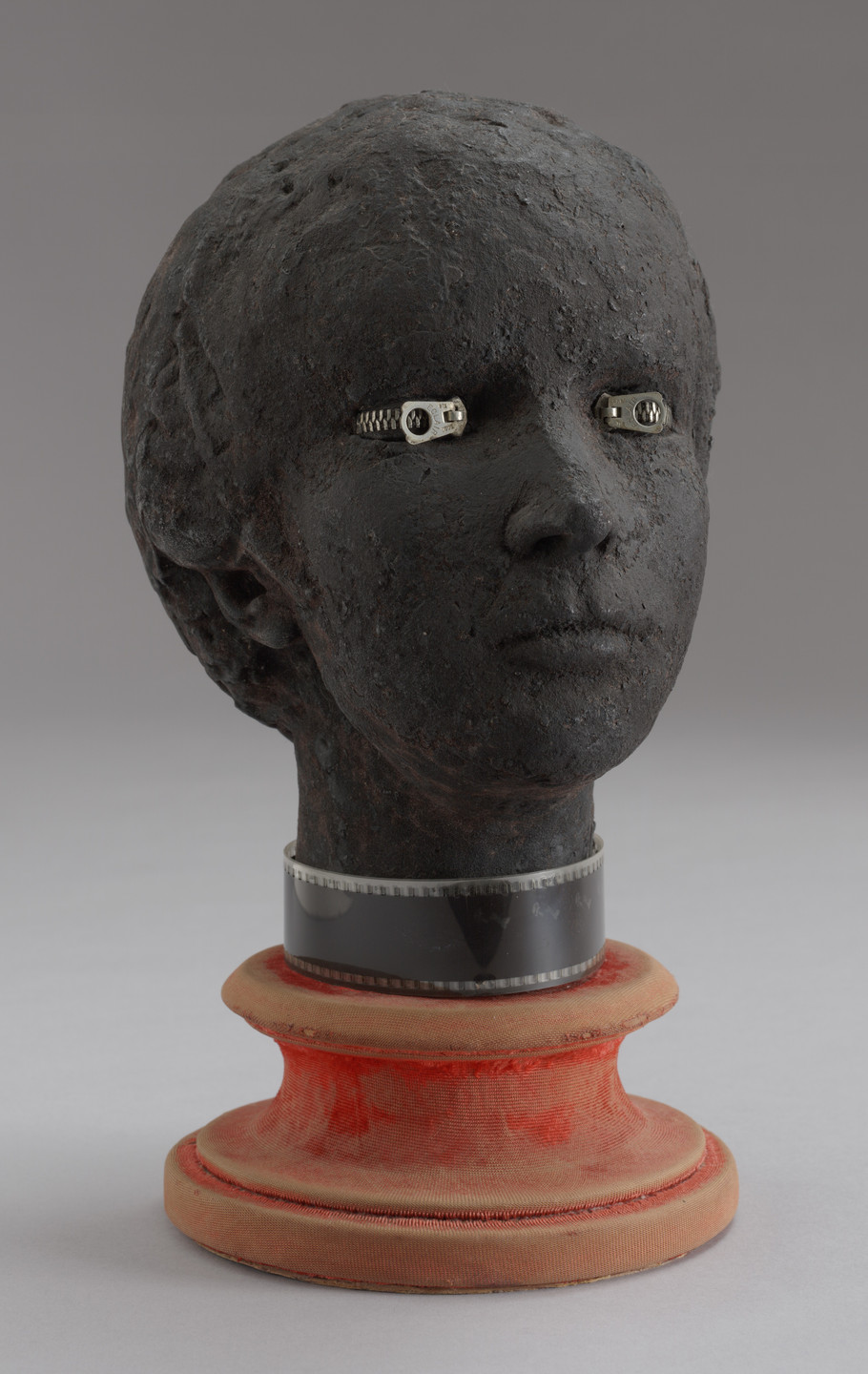 A dark, textured bust with metallic zippers for eyes and a filmstrip around the neck is perched on a distressed, red spool-shaped base. The background is plain gray. This is an AI generated description. To give feedback, email ai@moma.org.