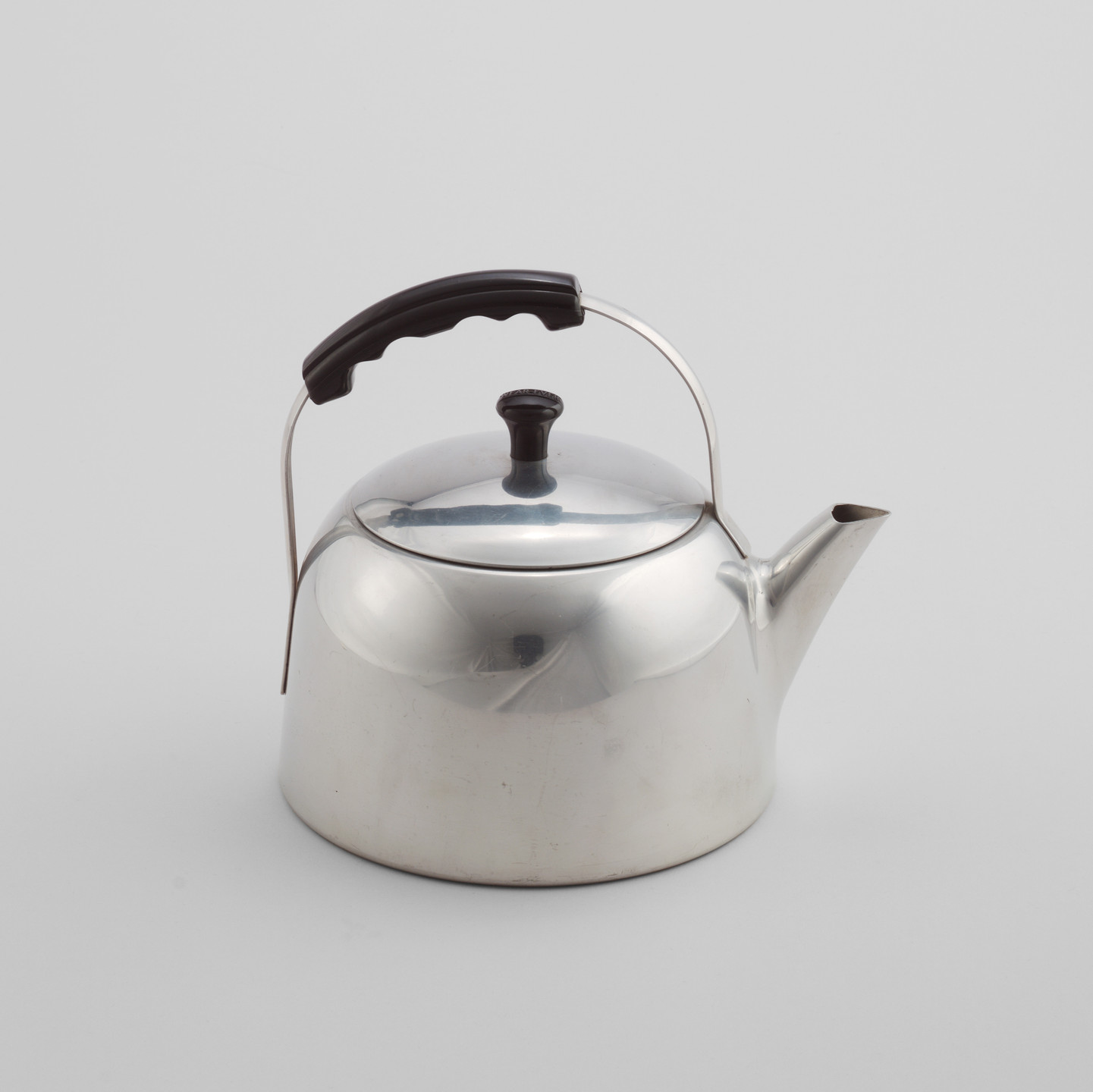 A silver metallic teapot with a black handle and knob is positioned against a plain, light grey background. The teapot has a smooth, rounded body and a short spout. This is an AI generated description. To give feedback, email ai@moma.org.