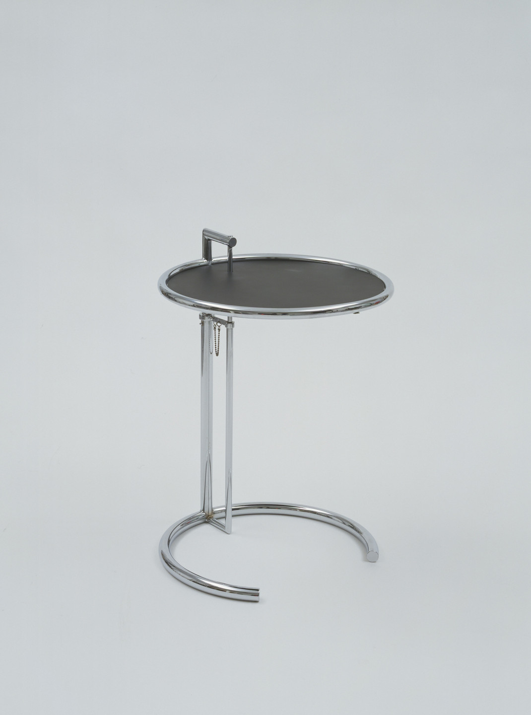 A small, round, black-topped table with a chrome frame and a semi-circular base stands against a plain light background. A vertical rod with a handle is attached to the top edge of the table. This is an AI generated description. To give feedback, email ai@moma.org.