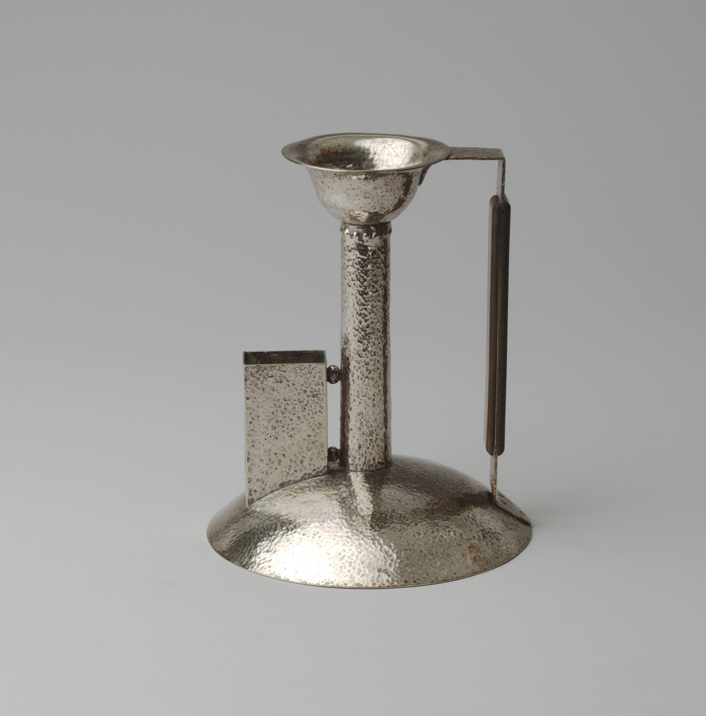 A silver colored metal object with a hammered texture featuring a circle base, a vertical cylindrical stem, and a shallow bowl-like top. It has a thin, vertical handle attached to the right side and a vertical rectangular form coming out of the left side of the base. This is an AI generated description. To give feedback, email ai@moma.org.