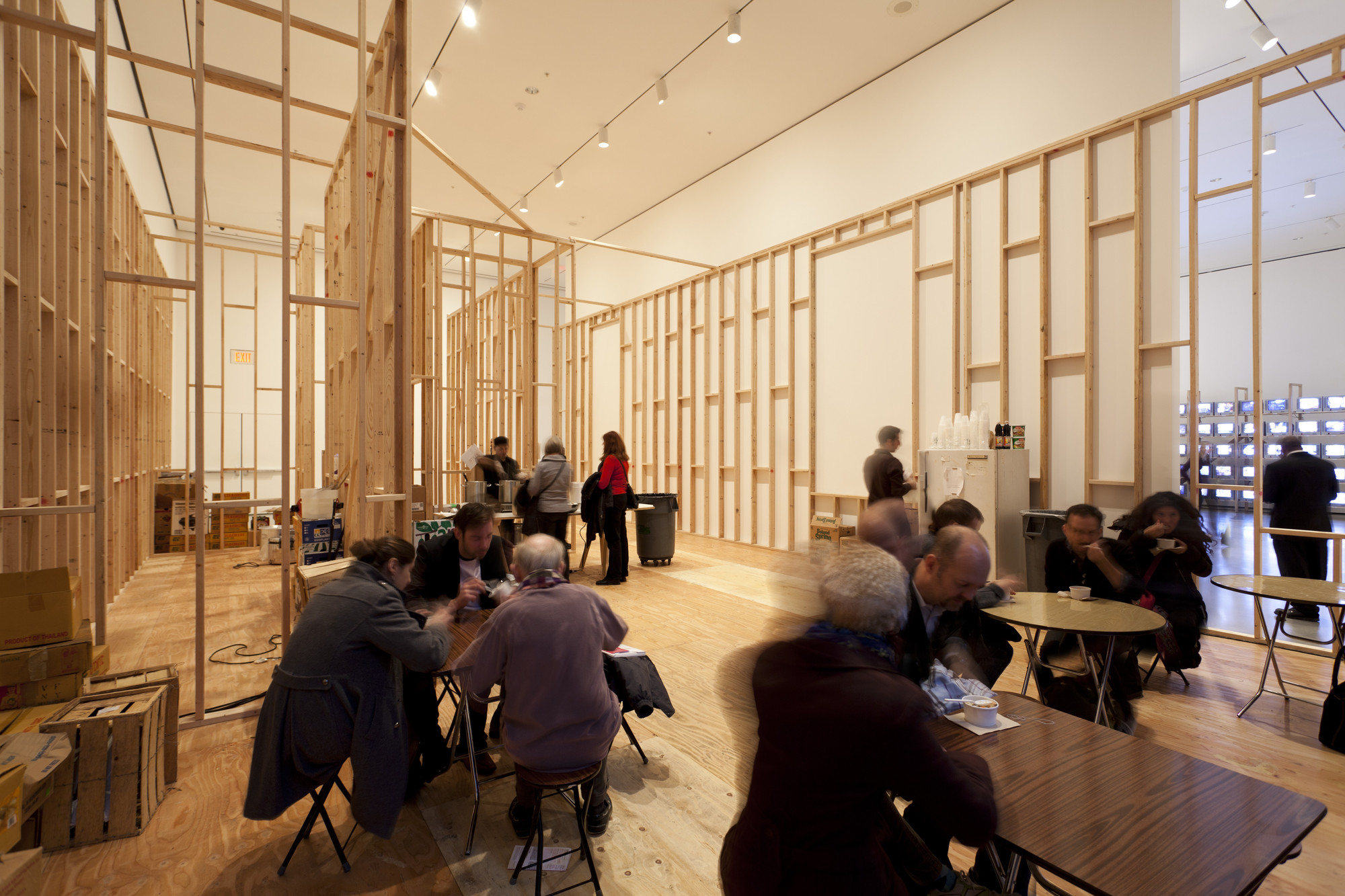 A spacious room with exposed wooden framing and light-colored walls, filled with several groups of people sitting at small tables and engaged in conversation. The lighting is bright, and the floor is made of wood. This is an AI generated description. To give feedback, email ai@moma.org.