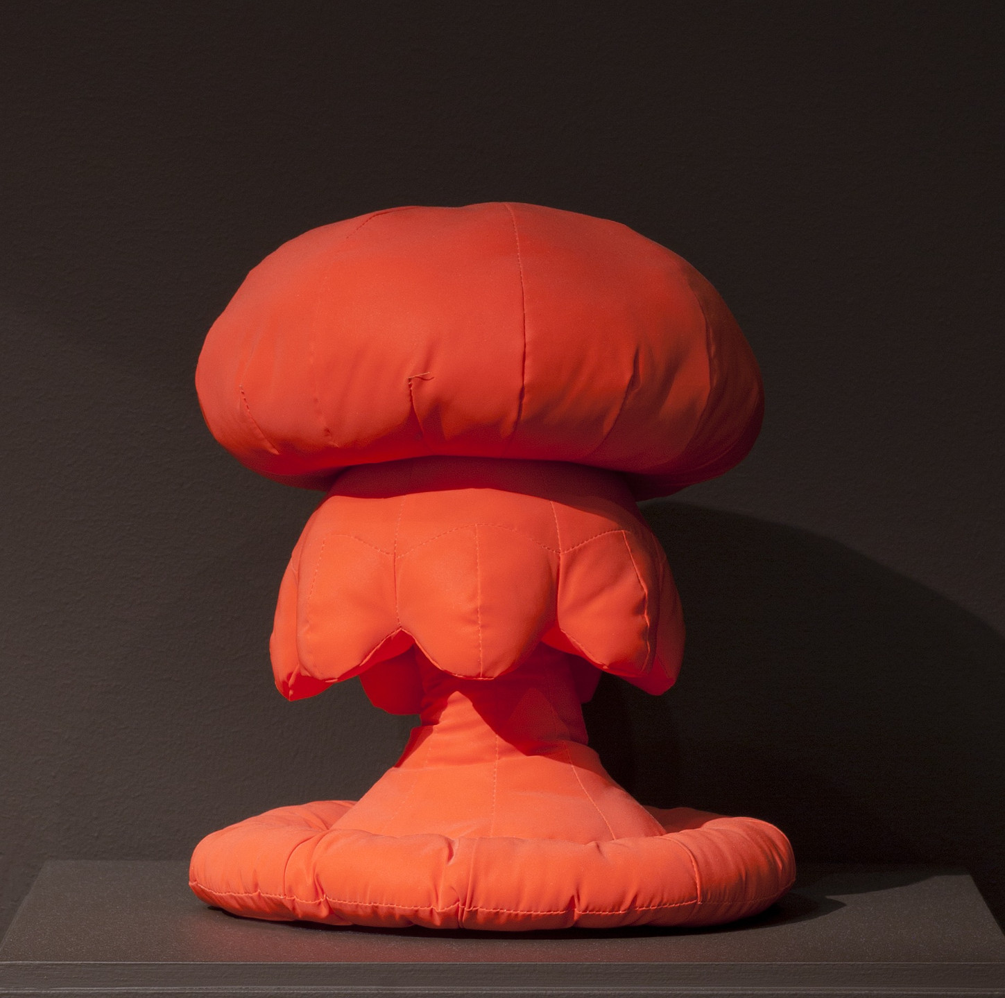 A bright orange, padded fabric sculpture on a dark background, shaped like a mushroom with a bulbous top and layered, petal-like sections. The sculpture stands on a round, cushioned base. This is an AI generated description. To give feedback, email ai@moma.org.