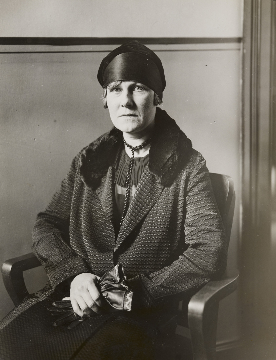 A person is seated in a chair and wearing a textured coat with a fur collar, a dark headscarf, and gloves. The image is in black and white with a neutral background. This is an AI generated description. To give feedback, email ai@moma.org.