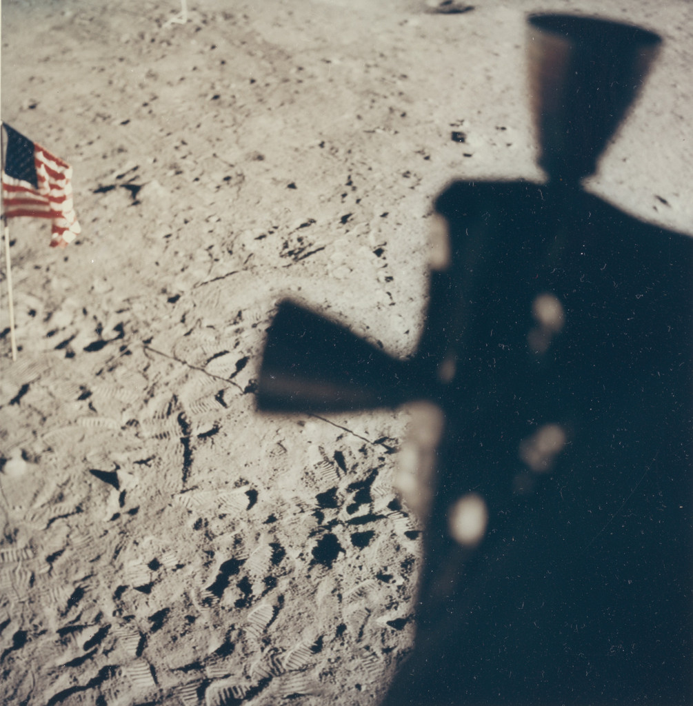 The surface of the moon. Gray dust, pockmarked and ancient. An astronaut's shadow stretches across the frame, distorted and ghostly. In the upper left corner, an American flag. Neil Armstrong or Buzz Aldrin, July 20, 1969.