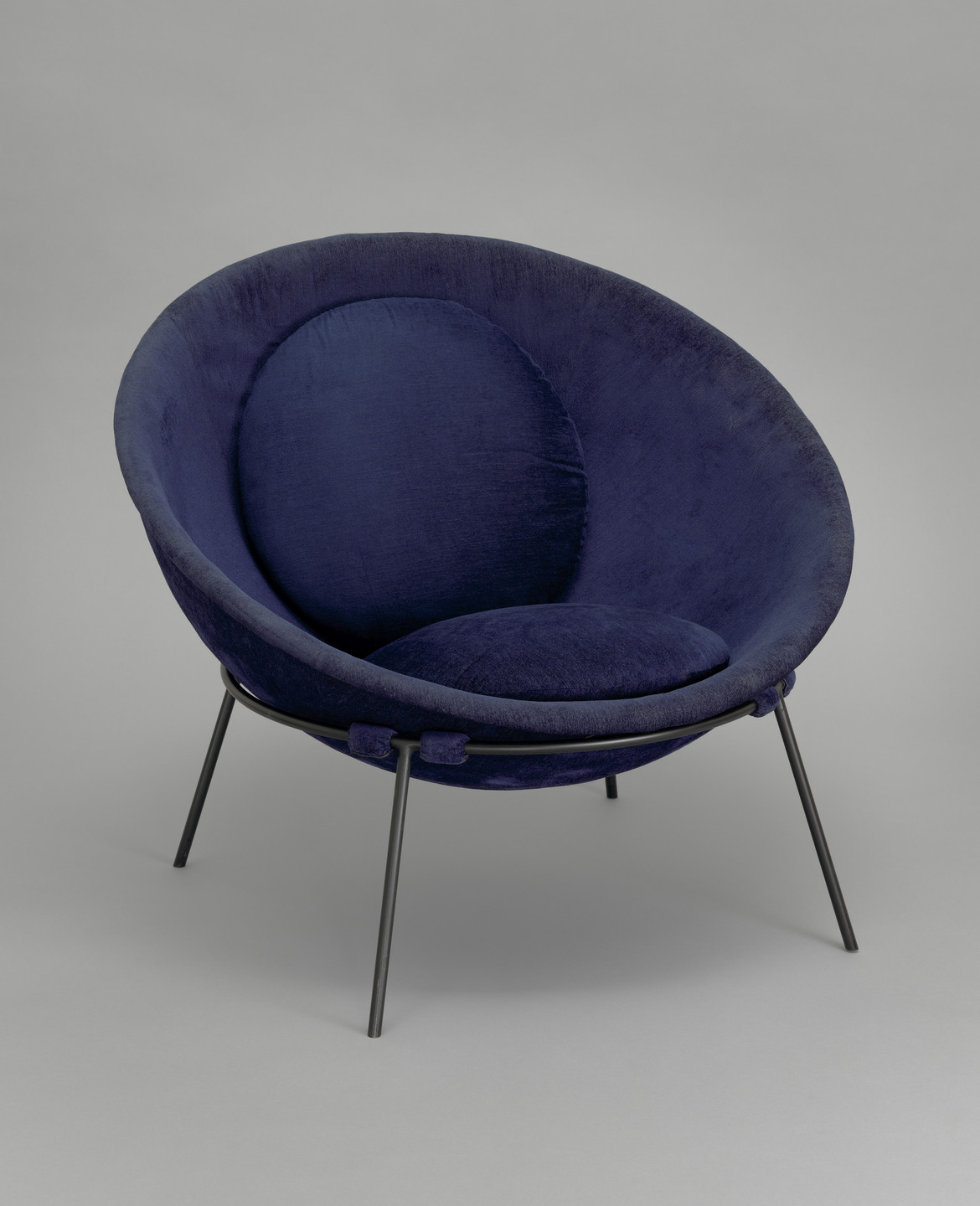 A deep blue, round chair with a padded seat and backrest is set against a plain grey background. The chair has thin, black metal legs extending outward. This is an AI generated description. To give feedback, email ai@moma.org.