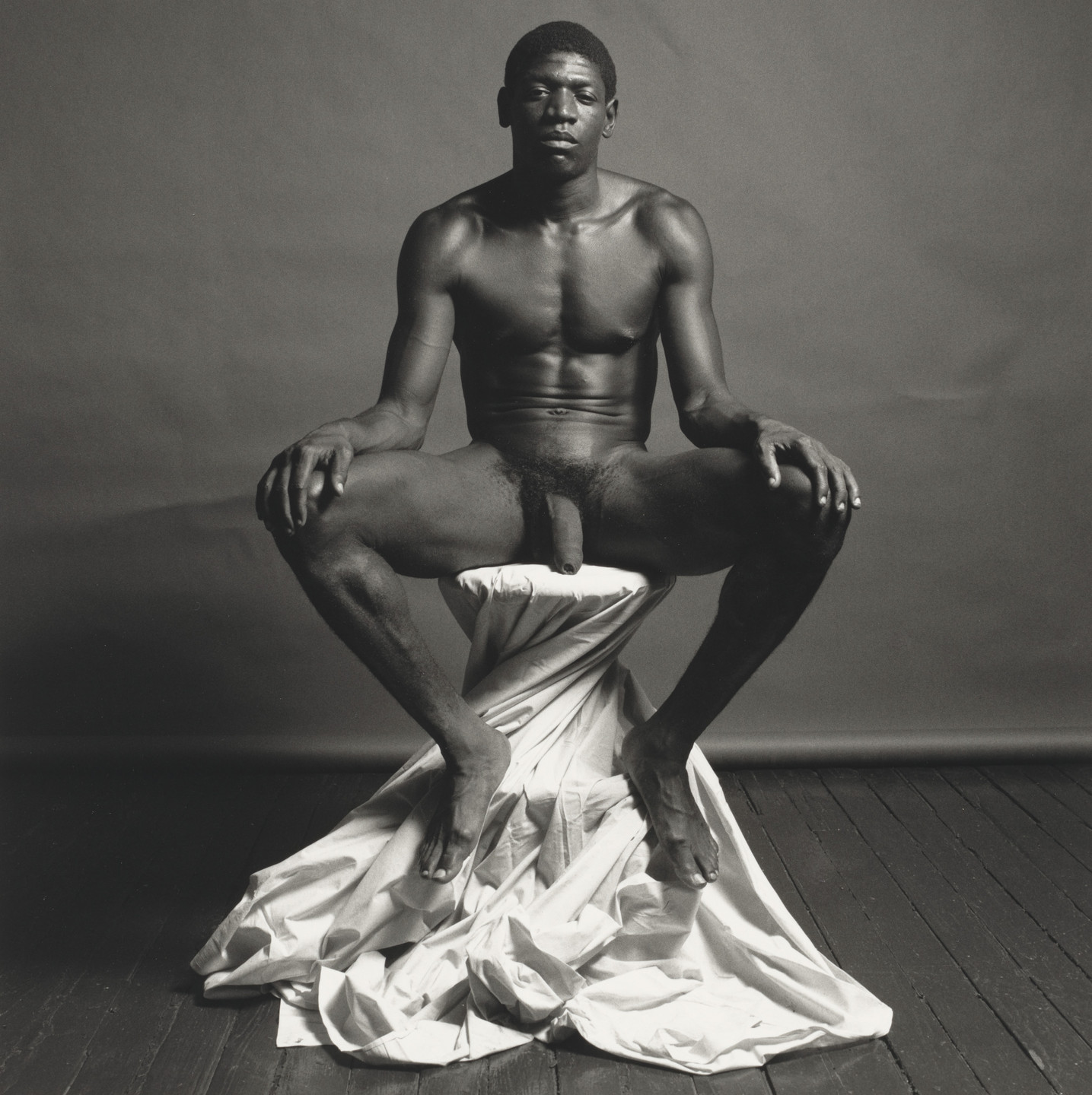 A person sits nude on a draped stool against a plain, dark background with a wooden floor. The image is in black and white with the person's legs spread wide. This is an AI generated description. To give feedback, email ai@moma.org.