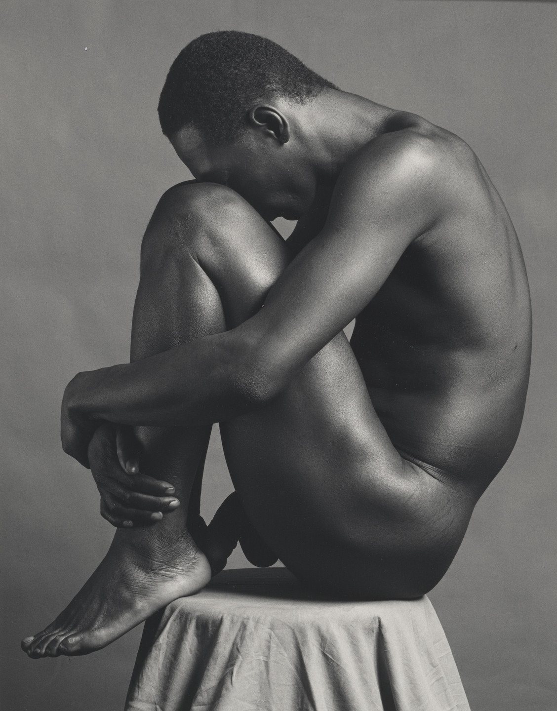 A nude person is sitting on a stool, hugging their knees and resting their head on them, with their face turned towards the right. The image is in grayscale, emphasizing the contours and the texture of their skin. This is an AI generated description. To give feedback, email ai@moma.org.