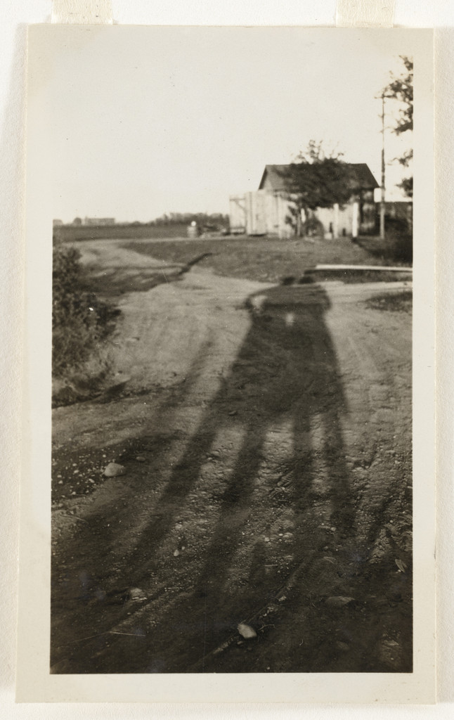 A small, vintage gelatin silver photograph showing a long shadow of a person stretched across a dirt road. The shadow reaches toward a small farmhouse in the distance. The photographer is invisible except for this dark silhouette cast by the sun.