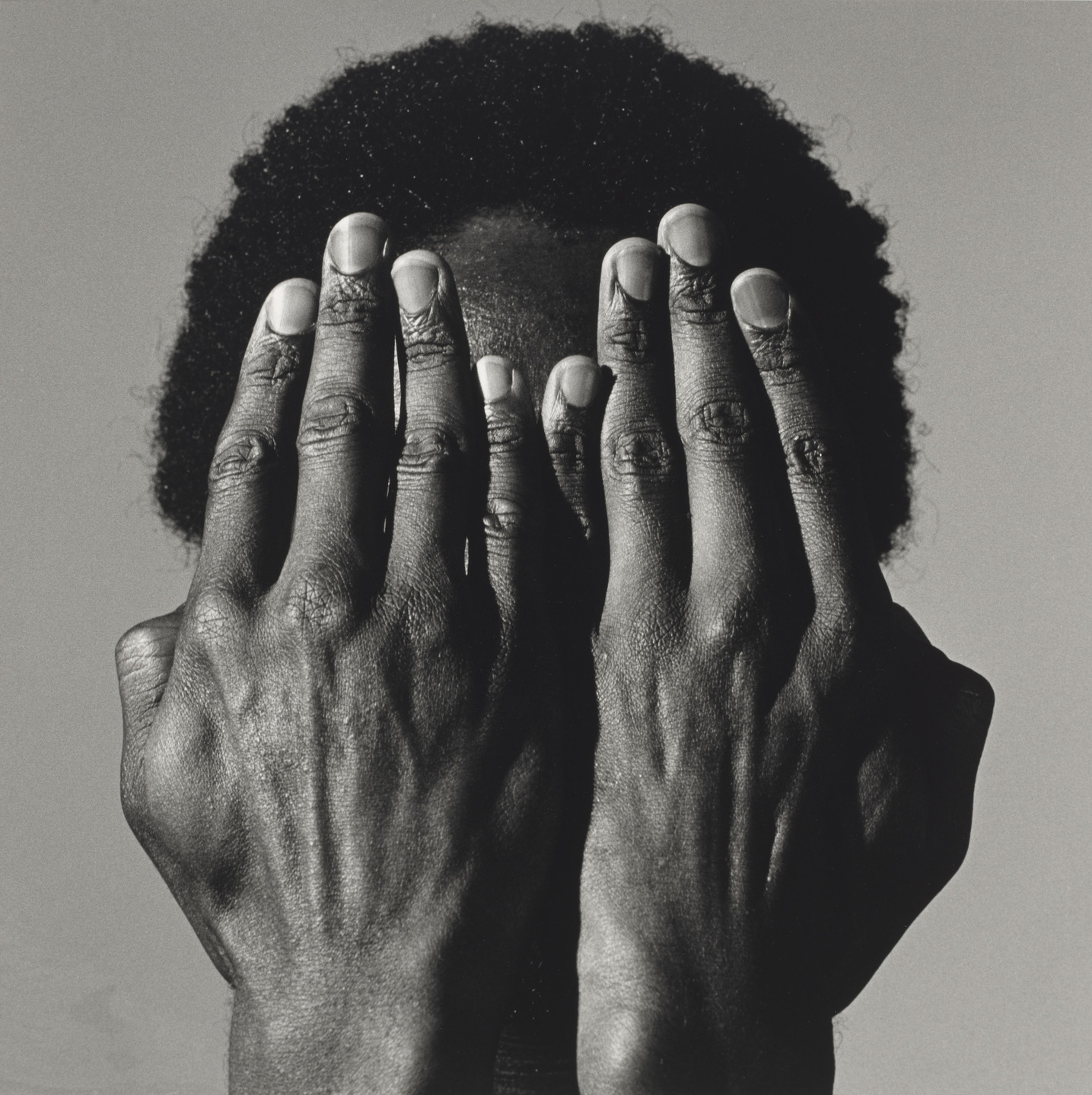 A person with short, curly hair covers their face with both hands, fingers slightly spread. The image is in black and white, emphasizing the texture of the skin and hair. This is an AI generated description. To give feedback, email ai@moma.org.