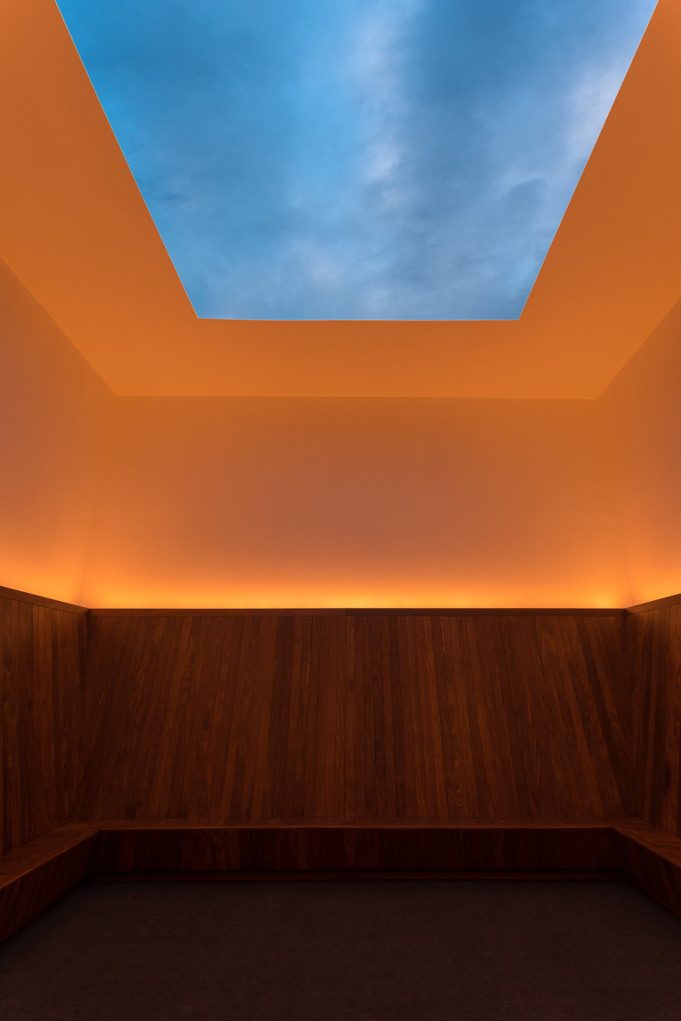 A room with wooden paneling and a ceiling featuring a large rectangular opening to the blue sky. Soft orange lighting illuminates the upper walls and ceiling edges. This is an AI generated description. To give feedback, email ai@moma.org.
