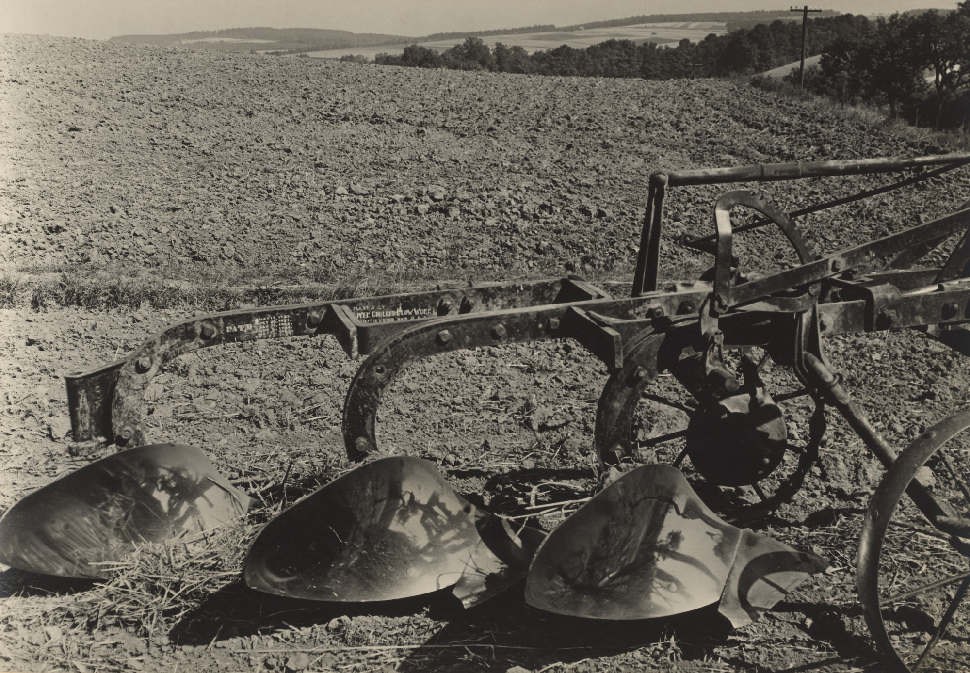 A black and white photo shows a metal plow in the foreground, set on a barren, freshly plowed field. In the distance, there are rolling hills and trees under a clear sky. This is an AI generated description. To give feedback, email ai@moma.org.
