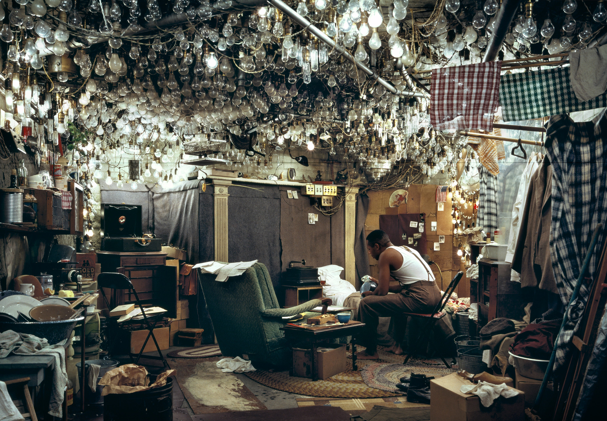 A room crowded with furniture, clothes, and hundreds of light bulbs hanging from the ceiling. The colors are predominantly earthy tones with some plaid fabric and a green armchair. This is an AI generated description. To give feedback, email ai@moma.org.