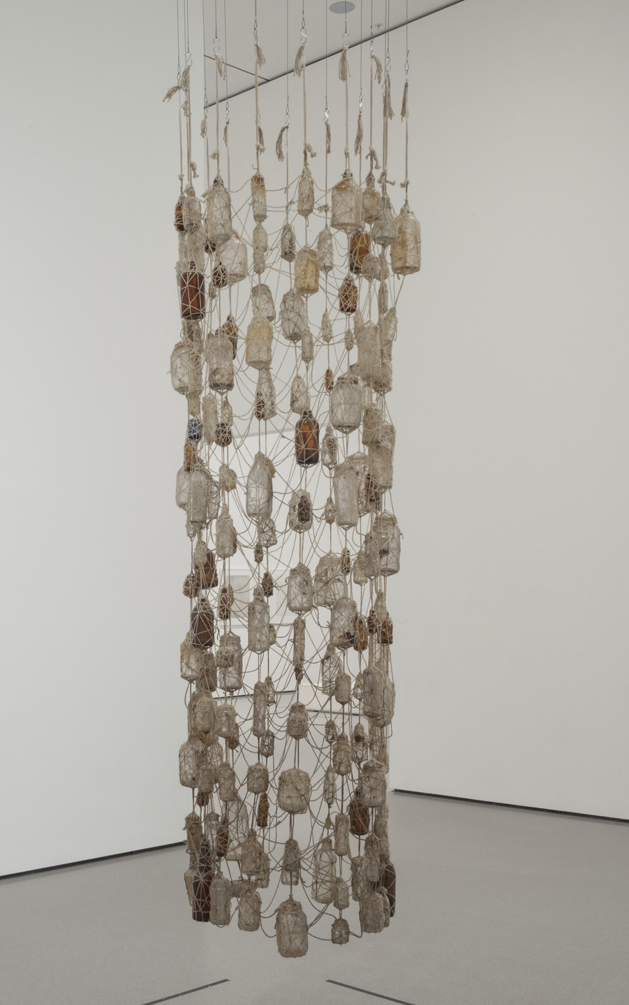 A cylindrical hanging sculpture composed of numerous small, beige, and brown objects encased in netting, suspended from the ceiling by thin strings. The background is a plain white wall. This is an AI generated description. To give feedback, email ai@moma.org.