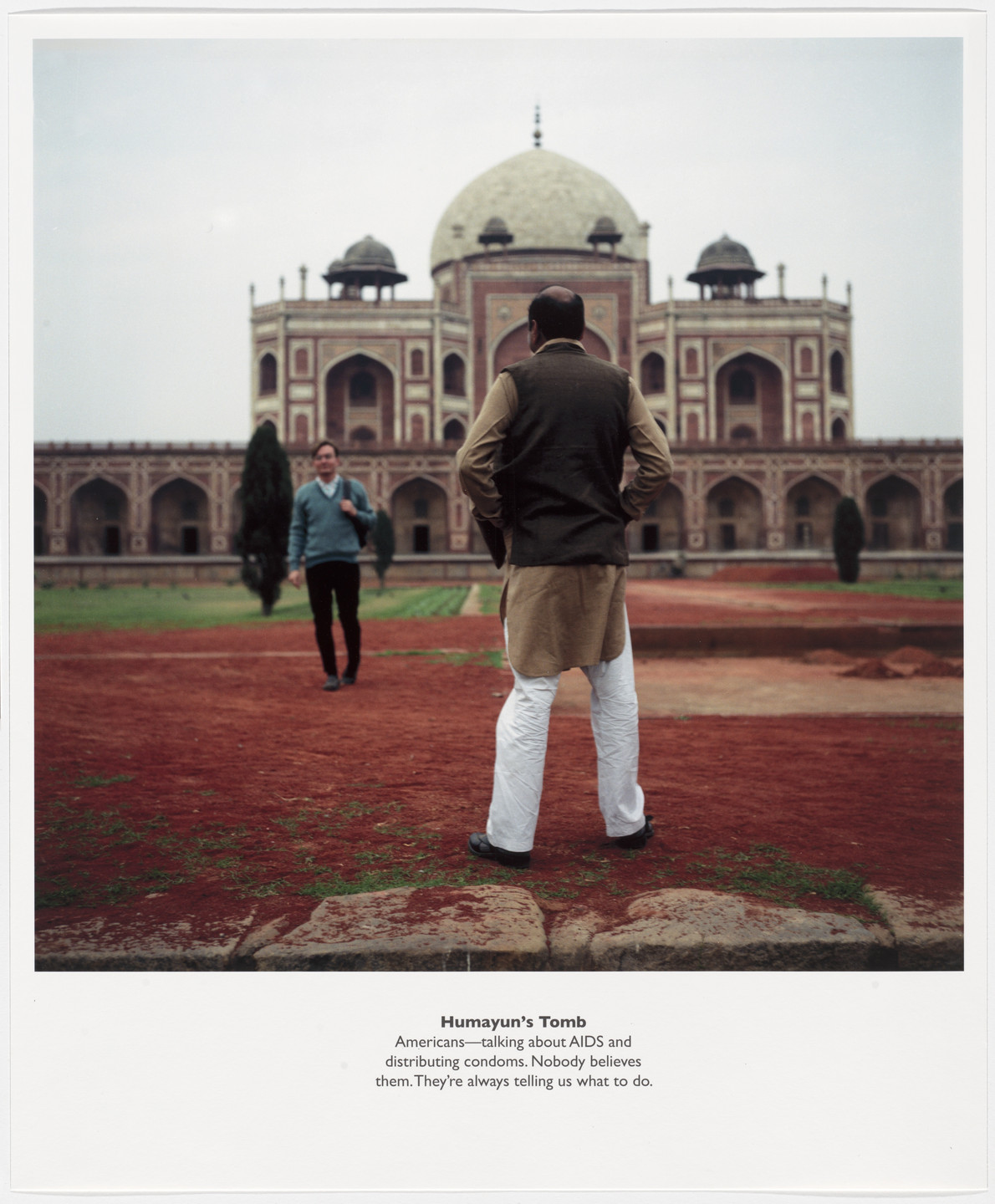 Two people are near a large, historic building with intricate archways and a domed roof, made of red and white materials. The foreground shows a reddish-brown dirt path with green patches of grass. This is an AI generated description. To give feedback, email ai@moma.org.